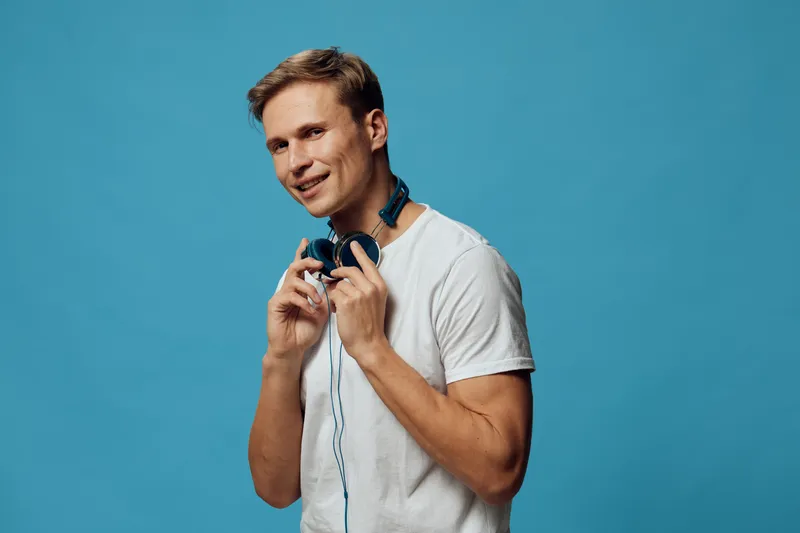Smiling young man with headphones holding headphones around his neck, wearing casual t-shirt, cheerful expression, happy mood, isolated on blue background, studio portrait, lifestyle concept.