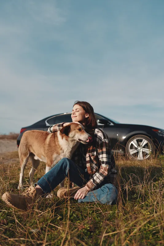 Woman sits in a sunlit field with her dog, enjoying a peaceful outdoors moment beside a sleek car, wearing a plaid shirt and denim jeans among tall grass and blue sky.