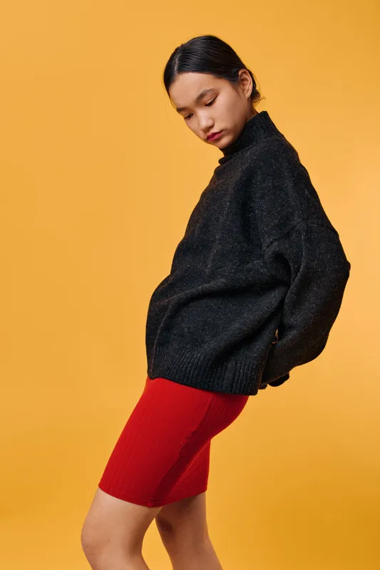 Woman wearing black sweater and red skirt standing against orange background in casual style. Young female model posing with head tilted and eyes closed for fashion portrait.
