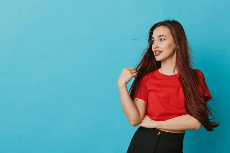 Young woman with long hair wearing red shirt and black pants posing confidently against blue background. Casual style, smiling face and natural look with hand near hair.