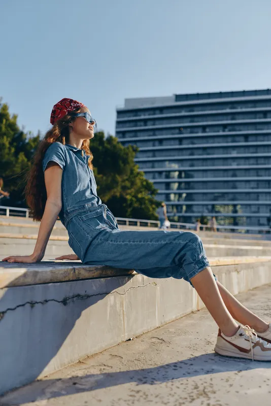 relaxed young woman sitting outdoors in casual denim outfit wearing sunglasses and red bandana enjoying sunny day near urban architecture