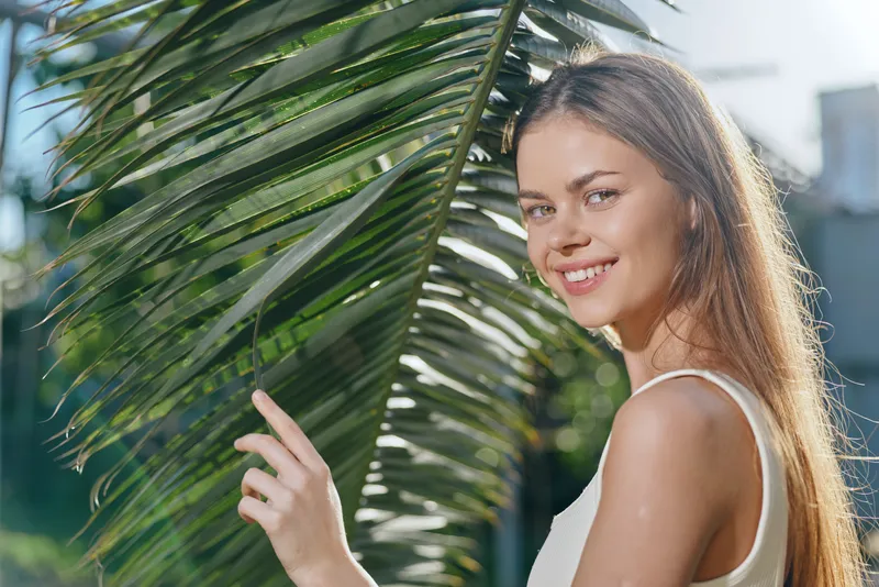 smiling woman touching palm leaf outdoors, natural light, casual white top, tropical greenery background, sunny day, joyful expression, young adult