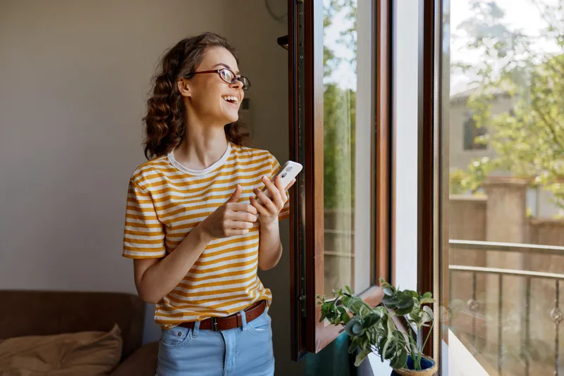 Young woman in a striped t shirt stands by a sunlit window smiling at her phone, enjoying a casual home moment in warm natural light
