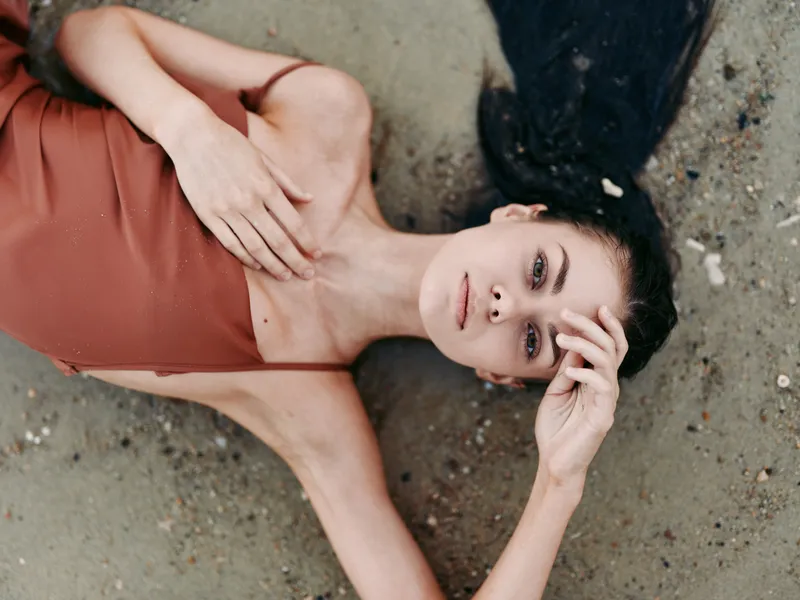 A woman lies on a sandy beach in a rust brown swimsuit, gazing softly at the camera with hair spread along the shore, evoking calm, warmth, and sunlit serenity.