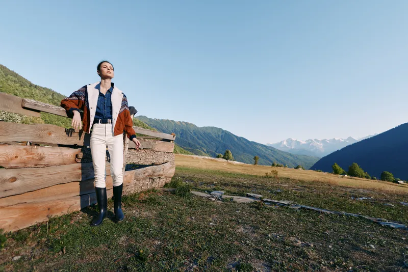 Woman stands in a mountains meadow leaning on a wooden fence wearing a stylish jacket and boots, bathed in warm sunlight across a wide landscape for travel and lifestyle.