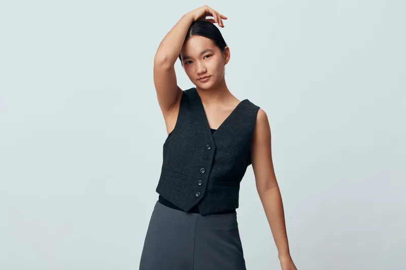 Young woman posing with arm raised and hand on head wearing sleeveless vest and skirt against plain light background. Fashion, casual style, portrait and confident expression.