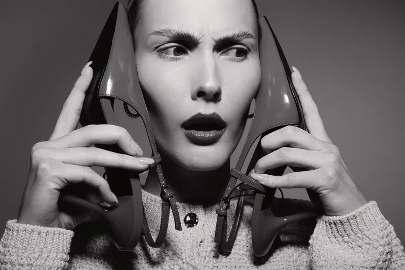 Confused Caucasian woman with light skin holds two shoes near her ears, wearing cozy sweater. Studio portrait in black and white, concept for confusion, fashion, and communication.