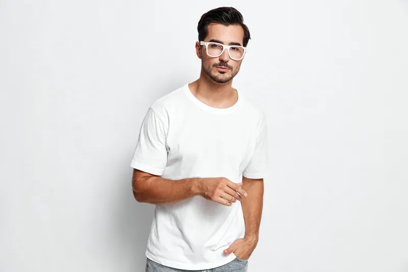 Young smiling man in casual white t-shirt and glasses standing against a light grey background, looking at the camera with a confident smile. Modern lifestyle concept.