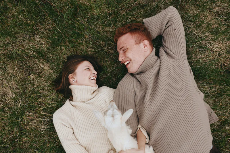 Cheerful couple outdoors, lying on grass, cozy sweaters, smiling at each other, playful pet nearby, sunlit moment, relaxed pose, candid lifestyle, natural colors, gentle breeze, warm connection