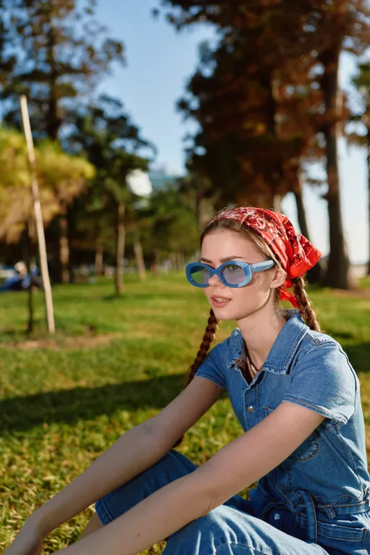 Lifestyle woman in modern boho-western denim street style sitting outdoors wearing blue sunglasses and a red bandana, captured with warm film color tones.