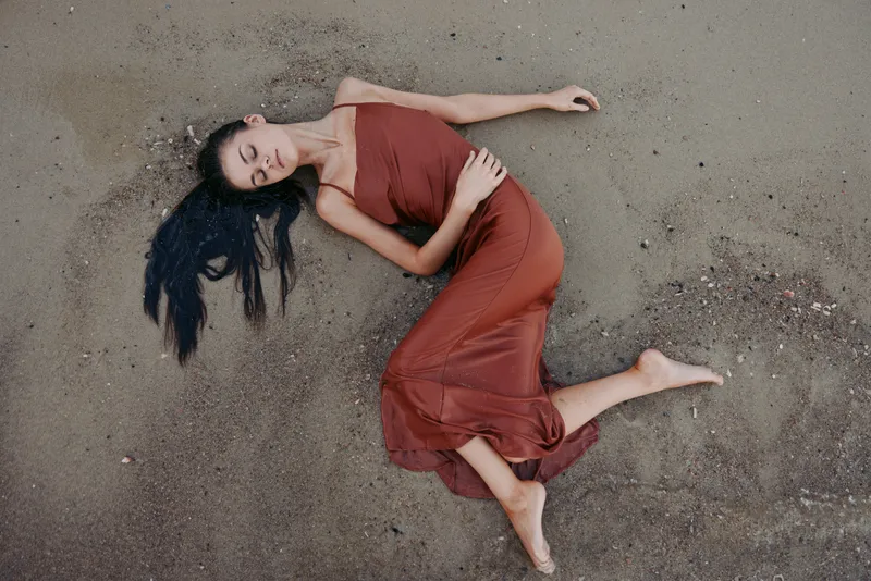A woman in a flowing red dress lies on dusty ground, eyes closed, gracefully posed with relaxed posture, soft light highlights fabric drape, texture, warmth, and sculpted silhouette.