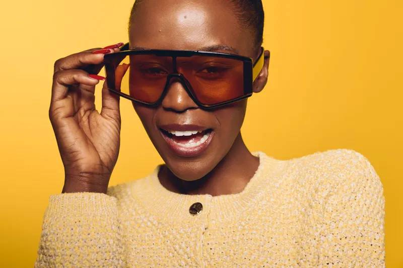 Close-up of a joyful black African woman with dark skin wearing oversized tinted sunglasses and a yellow knitted sweater against a bright yellow background. Concept of fashion and happiness.