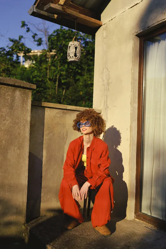 A stylish woman in a red outfit sits on a sunlit balcony, exuding casual elegance. A lifestyle portrait capturing confidence, warmth, and outdoor fashion