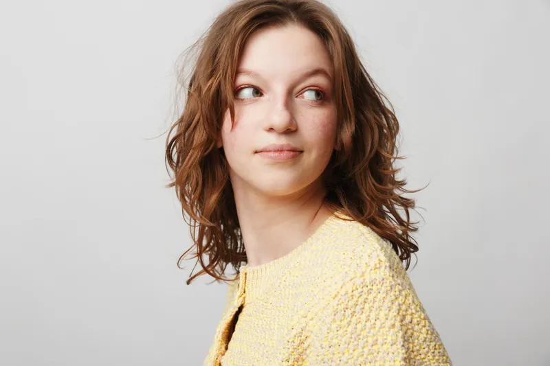 Young woman with curly hair wearing yellow sweater looks sideways with a thoughtful expression on a plain light background. Concept of casual style and natural beauty.