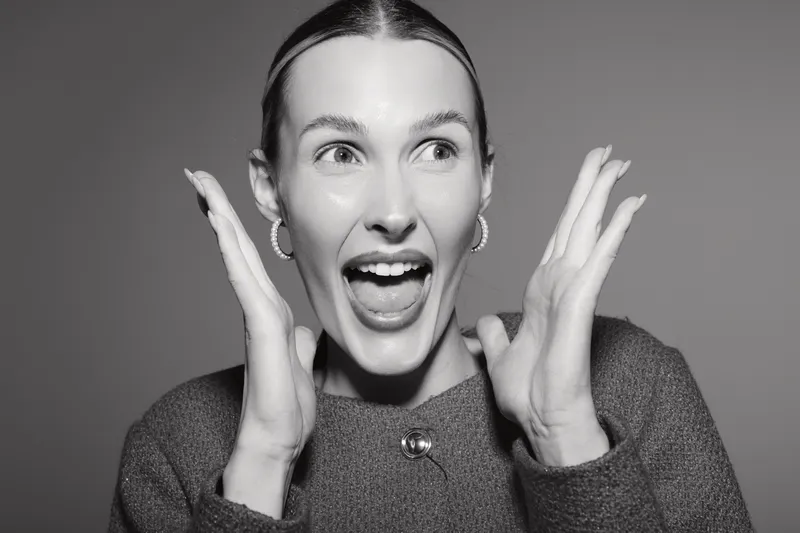 Excited Caucasian woman with fair skin and earrings expresses joy with open mouth and raised hands in black and white portrait.