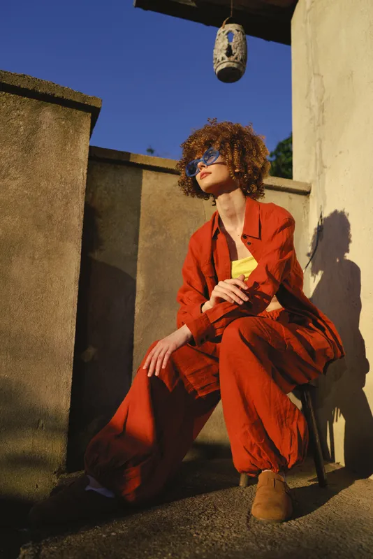 Vibrant woman outdoors in a burnt orange suit, seated on a terrace, sunlit moment, relaxed pose, sunglasses, curly hair, holding a yellow item, warm earthy tones