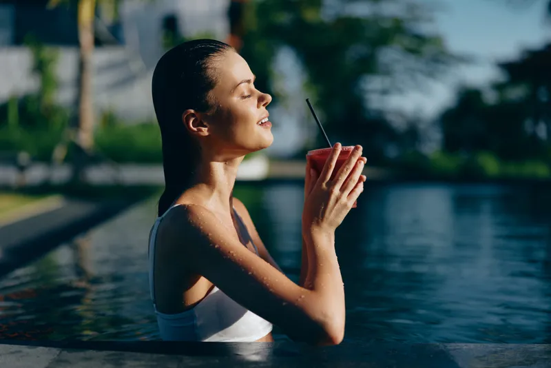 Bright, serene woman enjoys a quiet moment in a pool, standing waist deep and lifting a twig as a playful prop, expressing calmness, balance, and a healthy lifestyle