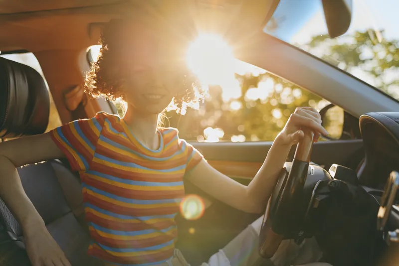 Joyful woman in a car, sunlit moment, striped shirt, relaxed posture, warm glow, casual drive, nature visible through window, goldenhour atmosphere