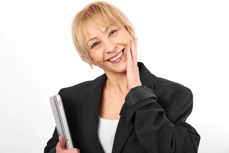 Woman smiling while holding smartphone, blonde hair and blazer, portrait on white background, studio lighting, showing communication and technology for senior confidence