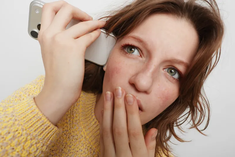 Young woman with red hair and freckles holds smartphone to ear and covers mouth with hand. She looks surprised or shocked against plain white background. Concept of communication and emotion.