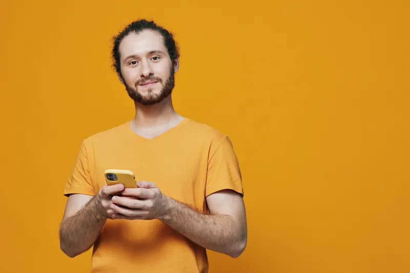 Cheerful young man using smartphone against a bright yellow background, dressed in a casual orange t shirt, embodying positivity and modern technology