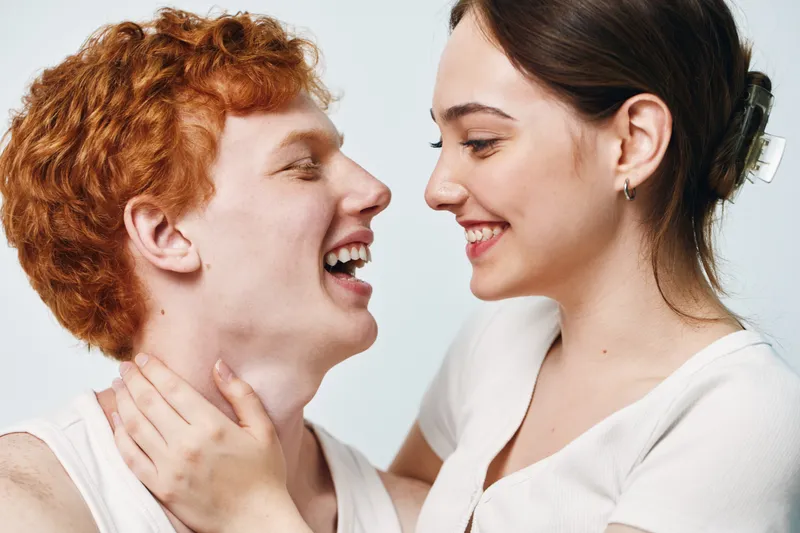 Young couple smiling and embracing in casual clothes against white background. Man and woman looking at each other with happiness and affection in closeup portrait.