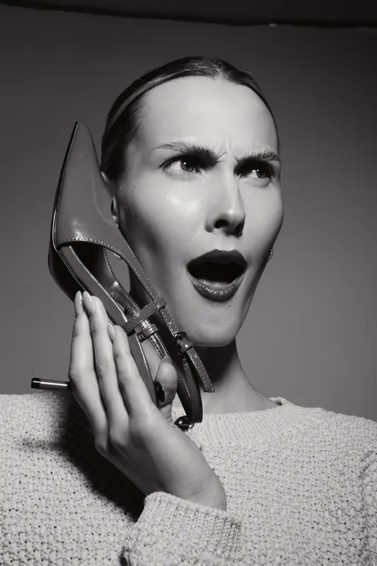 Caucasian woman with fair skin holds high heel shoe to her ear with confused expression. Black and white studio portrait with dramatic lighting and textured sweater. Concept of surprise and emotion.