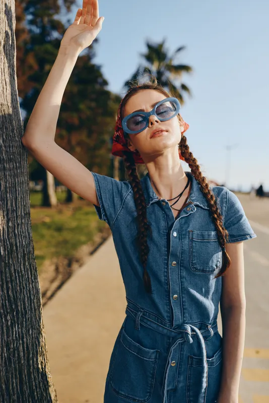 Lifestyle woman in modern boho-western denim street style with film color effects, wearing sunglasses and braids, posing outdoors on a sunny day.