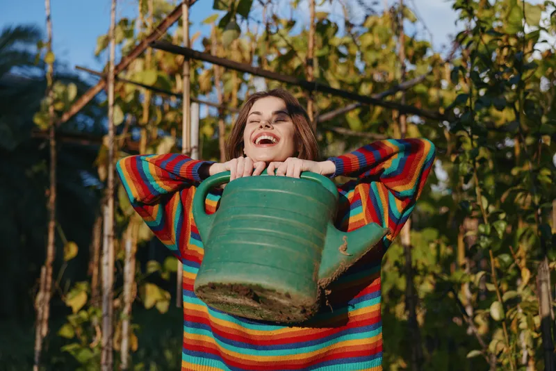Woman holding watering can in garden, smile bright as she wears a colorful striped sweater outdoors among plants and trellis, sunlit portrait of joyful gardening and leisure.