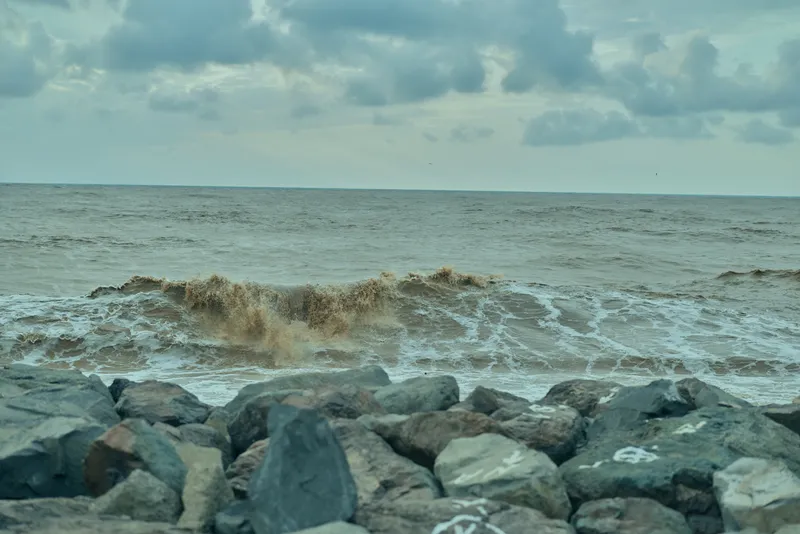 Coastline with rugged rocks along the shore as waves crash under a cloudy sky, sea spray in the air, and a moody seascape perfect for nature, travel, and outdoor weather scenes