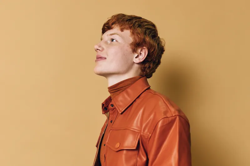 Young man with red hair wearing orange leather jacket and turtleneck looking up thoughtful against beige background in studio portrait.