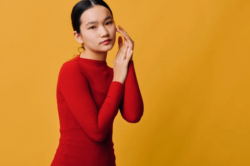 Young woman in red sweater posing and touching face with hands looking at camera on yellow background. Studio portrait with confident expression and stylish casual clothing for fashion.