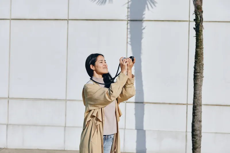 photography, woman, camera, outdoors, daylight, lens, pose, fashion, sunlight, wall A stylish woman in a beige trench stands before a white tiled wall, lifting her camera to capture a bright day with