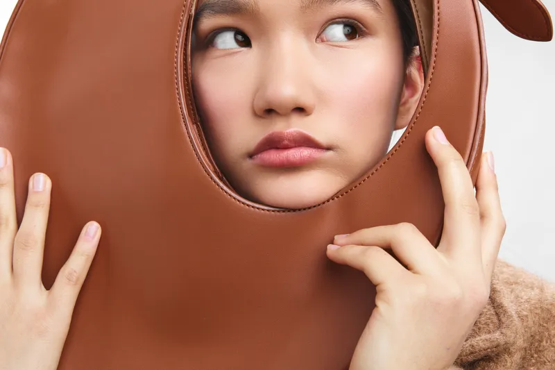 asian woman dons a large brown leather mask in a studio portrait, her eyes glancing aside with a thoughtful, curious smile that adds a playful, enigmatic vibe.