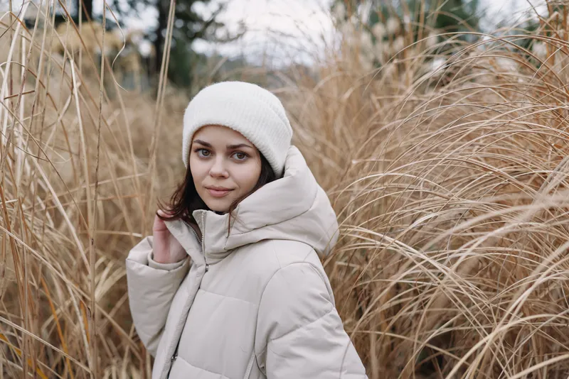 Winter portrait of a woman in a light coat and beanie among tall grasses, outdoors, calm mood, soft natural light, seasonal fashion style and cozy comfort