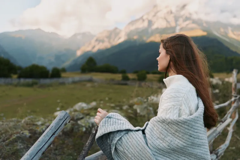 A young woman sits on a wooden fence in a mountain valley, wrapped in a chunky knit sweater, gazing into the distance with a calm, contemplative mood.