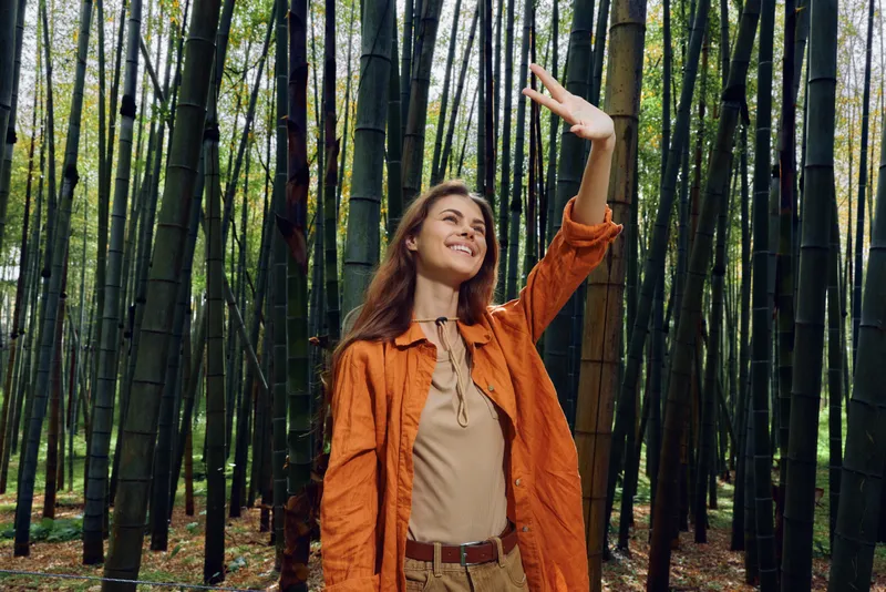 Woman in orange jacket smiles and explores a dense bamboo forest outdoors, nature portrait showing walking leisure, joy and curiosity among tall trees with dappled sunlight.
