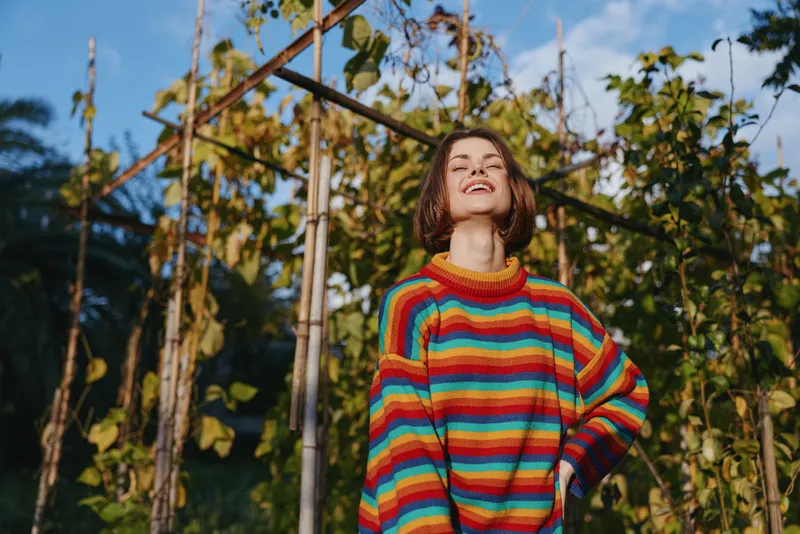 Young adult woman smiling in a rainbow striped sweater in a tropical garden portrait, casual outfit, short hair, joyful mood, sunlight, lifestyle and fashion outdoor scene
