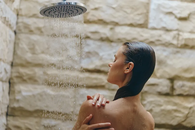 woman showering outdoors under rain shower with stone wall background, wet hair, relaxed mood, natural spa atmosphere, wellness and self-care moment