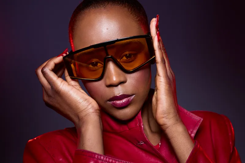 Portrait of a confident dark skinned African woman wearing large orange sunglasses and a red leather jacket. Studio lighting highlights her smooth skin and stylish look. Fashion concept.