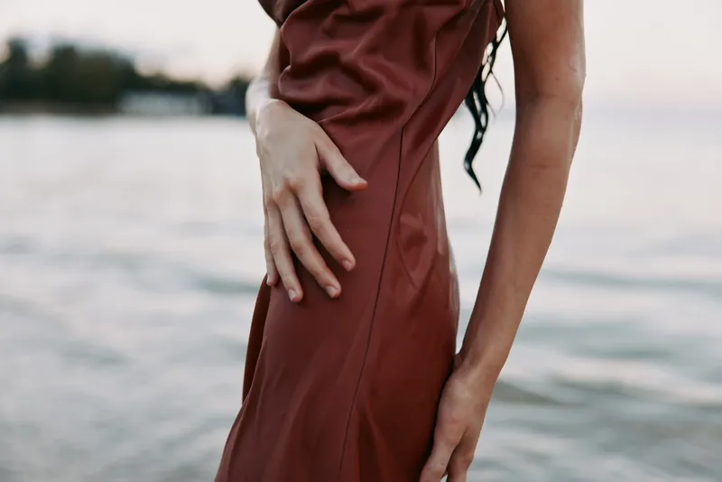 Close shot of a graceful torso and hand resting on a brown dress by a calm shoreline, focusing on texture, posture, and subtle elegance against soft water and horizon.