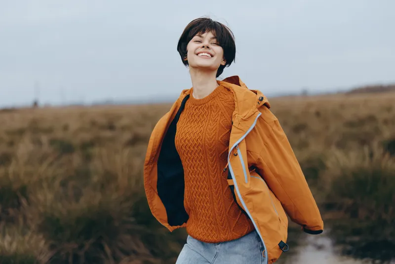 Woman outdoor lifestyle photo, smiling woman walking along a rural path in a field, wearing orange jacket and knitted sweater, cloudy sky, natural light open countryside landscape wide
