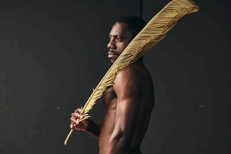 African man with dark skin holding large golden feather in creative conceptual visual style. Portrait, artistic expression and cultural identity with strong body and serious look on dark background.