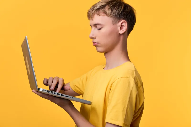 Boy in a yellow shirt holds a laptop, focused on the screen, showcasing technology, learning, and online work in a bright studio setting for stock use.
