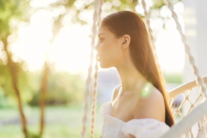 Young woman in white dress with long hair, enjoying sunlight on a swing in nature, conveying serenity and beauty in a bright, warm setting
