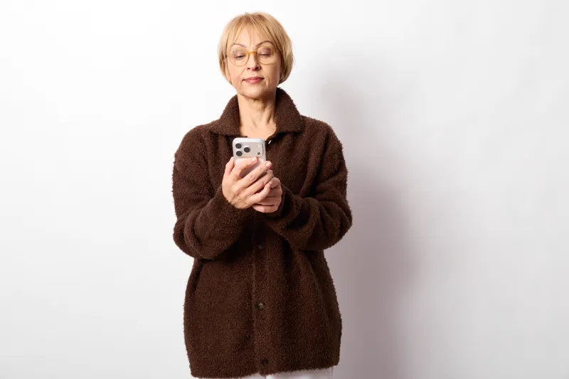 Senior woman with glasses holds smartphone for selfie in indoor scene, neutral expression about technology communication and privacy on plain white background