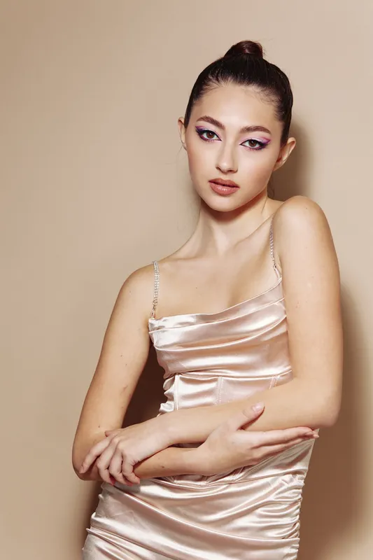 Young woman with sleek updo wears a silky light pink dress, posing with crossed arms on a neutral background. Creative makeup highlights sharp features, beauty and fashion concept.