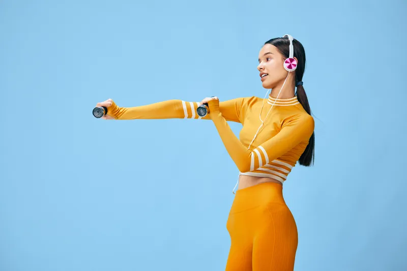 Young woman fitness workout in bright studio, wearing orange activewear and pink headphones, lifting small dumbbells in a dynamic pose against a smooth pale blue background.