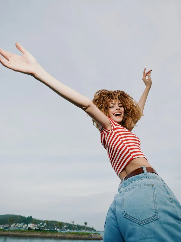 Joyful young woman in a striped red and white top and denim jeans spreads arms wide, jumping outdoors under a bright sky, conveying freedom, happiness, and carefree energy, for a vibrant lifestyle