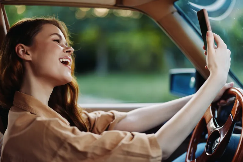 Young woman enjoying a joyful moment while driving in a vintage car, taking a selfie against a lush green background with natural light.